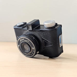 Könnte beinhalten: Eine schwarze Waldorf Minicam 35mm Filmkamera mit silbernem Objektiv und silbernem Oberknopf. Die Kamera hat einen schwarzen Lederriemen und eine silberne Metallplatte an der Seite.