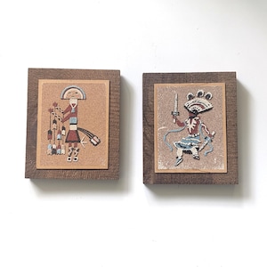 Puede incluir: Dos pinturas de arena nativas americanas enmarcadas. Una presenta una figura sosteniendo flechas, la otra una figura con una espada. Ambas están en marcos de madera con un fondo beige.