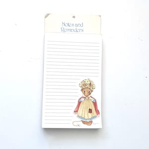 May include: A rectangular notepad with horizontal lines and the text Notes and Reminders printed at the top. The bottom right corner features a colorful illustration of a mouse wearing a yellow polka dot bonnet, a red shawl, and an apron.