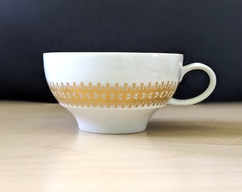 Peut inclure: Tasse blanche avec une bande décorative dorée. La tasse a une forme arrondie et une anse. La bande dorée présente un motif répété de formes stylisées. La tasse est posée sur un fond neutre.