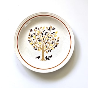 May include: A round, off-white ceramic plate with a brown rim and a tree design. The tree has brown branches, yellow and brown leaves, and black berries. Three black birds are at the base of the tree.