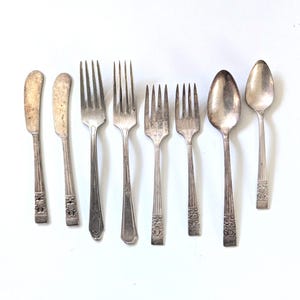 May include: An assortment of vintage silverware, including butter knives, forks, and spoons, arranged on a white surface. The silverware has a tarnished, antique appearance, with decorative floral patterns on the handles.