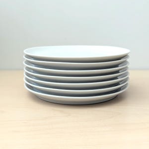 Pode incluir: Uma pilha de sete pratos redondos de cerâmica branca. Cada prato apresenta uma superfície lisa e plana com uma borda ligeiramente elevada. Os pratos são uniformes em tamanho e cor, empilhados verticalmente para criar uma aparência limpa.