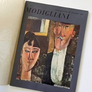 May include: A gray book cover with the title "MODIGLIANI" in white text. The Museum of Modern Art is printed in smaller white text below the title. The cover features a painting of a man and woman with a dark background.