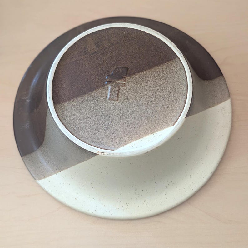 May include: A ceramic plate with a speckled, off-white rim and a color-blocked design. The plate's underside features a brown, tan, and off-white color scheme. A stamped mark is visible on the center of the plate. The plate is likely for serving food.