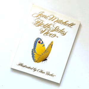 May include: A book cover with the title "Joni Mitchell Both Sides Now" in gold script. A yellow butterfly with blue and red accents is featured on the cover. The text "Illustrated by Alan Baker" is below the butterfly.