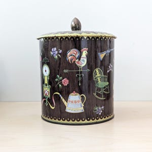 Puede incluir: Un bote de metal marrón vintage con tapa. El bote está decorado con ilustraciones coloridas de una veleta de gallo, flores, un reloj, una tetera y una mecedora. La tapa tiene un borde dorado.