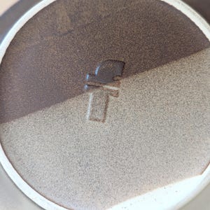 May include: Close-up of a ceramic plate with a two-tone brown and tan color scheme. The plate has a white rim and a stamped design in the center. The design appears to be a stylized letter or symbol.
