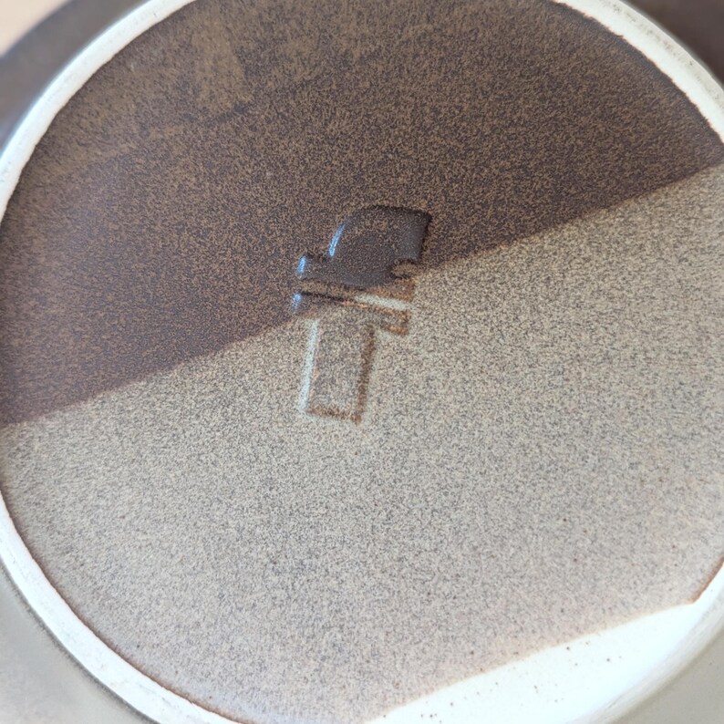 May include: Close-up of a ceramic plate with a two-tone brown and tan color scheme. The plate has a white rim and a stamped design in the center. The design appears to be a stylized letter or symbol.