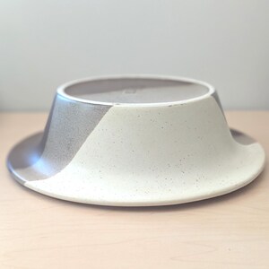 May include: A ceramic bowl with a wide, flat brim and a conical center. The bowl is divided into three sections: a light beige, a speckled white, and a gray section. The bowl is sitting on a light wood surface.