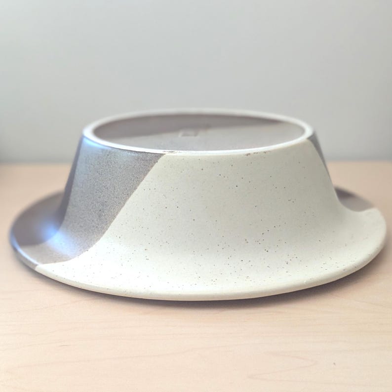 May include: A ceramic bowl with a wide, flat brim and a conical center. The bowl is divided into three sections: a light beige, a speckled white, and a gray section. The bowl is sitting on a light wood surface.