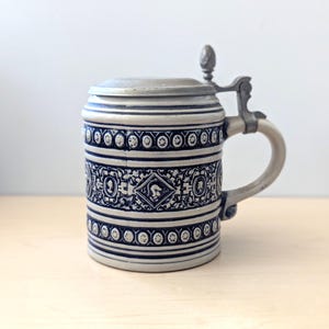 May include: A grey and cobalt blue ceramic beer stein featuring a metal hinged lid and handle. The body displays intricate embossed patterns, including floral motifs, geometric borders, and a central diamond shape with the text 15 56.