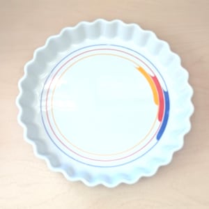 May include: White ceramic pie dish with a scalloped edge and a colorful rainbow design. The dish has a shallow, round shape and is suitable for baking and serving pies, tarts, or quiches. The dish is on a light wood surface.