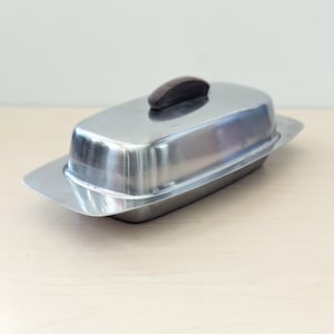 May include: A stainless steel butter dish with a rectangular shape and a dark brown wooden handle on the lid. The dish has a shiny, reflective surface and a slightly raised base. A classic kitchenware item.