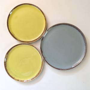 Puede incluir: Tres platos de cerámica en tonos amarillo y azul grisáceo. Los platos tienen forma redonda con un borde marrón. Los platos están dispuestos sobre una superficie blanca. Los platos son probablemente para servir comida.