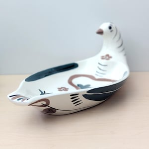 May include: A white ceramic dove-shaped dish with a raised head and tail. The dish is decorated with dark blue, brown, and black accents, including floral and geometric patterns. The dove's head is white with dark blue stripes.