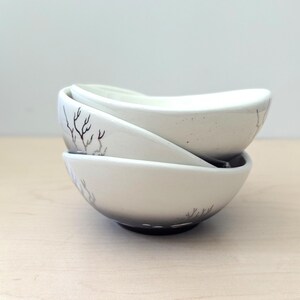 May include: Three off-white ceramic bowls stacked together. Each bowl has a unique, organic shape and is decorated with a delicate, hand-painted branch design in a muted brown color. The bowls are ideal for serving snacks or small portions.