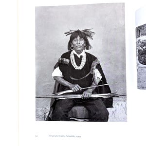 Może przedstawiać: Czarno-białe zdjęcie osoby w tradycyjnym stroju rdzennych Amerykanów. Osoba siedzi i trzyma łuk i strzałę. Osoba ma na sobie ciemną szal, biały naszyjnik i nakrycie głowy z piór. Obraz pochodzi z książki i zawiera tekst "Hopi portraits, Sekatila, 1901."