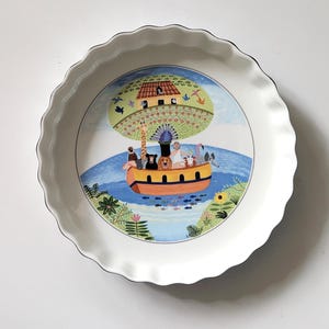 Może przedstawiać: Biała ceramiczna forma do ciasta z falistym brzegiem i czarnym obramowaniem. Środek przedstawia kolorową ilustrację Arki Noego z różnymi zwierzętami, domem i drzewem. Projekt zawiera niebieską wodę, zielone liście i niebieskie niebo.