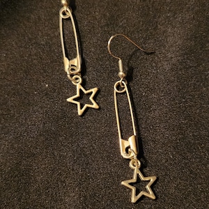 May include: A pair of gold-toned earrings featuring a safety pin design with a star charm dangling from the bottom.