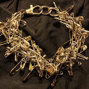 May include: A silver safety pin necklace made of many safety pins linked together. The necklace has a silver clasp.