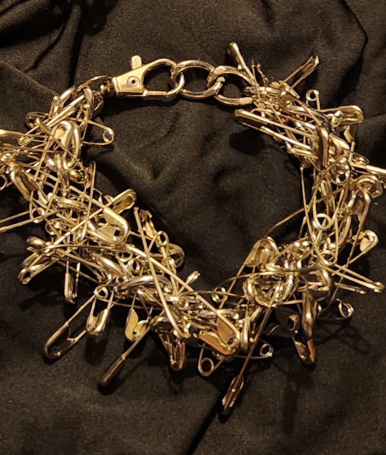 May include: A silver safety pin necklace made of many safety pins linked together. The necklace has a silver clasp.