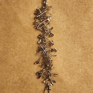 May include: A gold safety pin bracelet made of many safety pins linked together. The bracelet has a clasp closure.