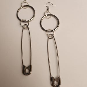 May include: A pair of silver earrings with a large circle and a safety pin dangling from each. The earrings are made of metal and have a simple design.