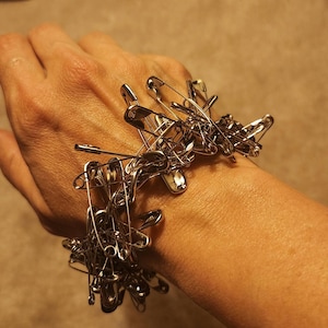 May include: A silver safety pin bracelet made of multiple safety pins linked together. The bracelet is worn on a person's wrist.