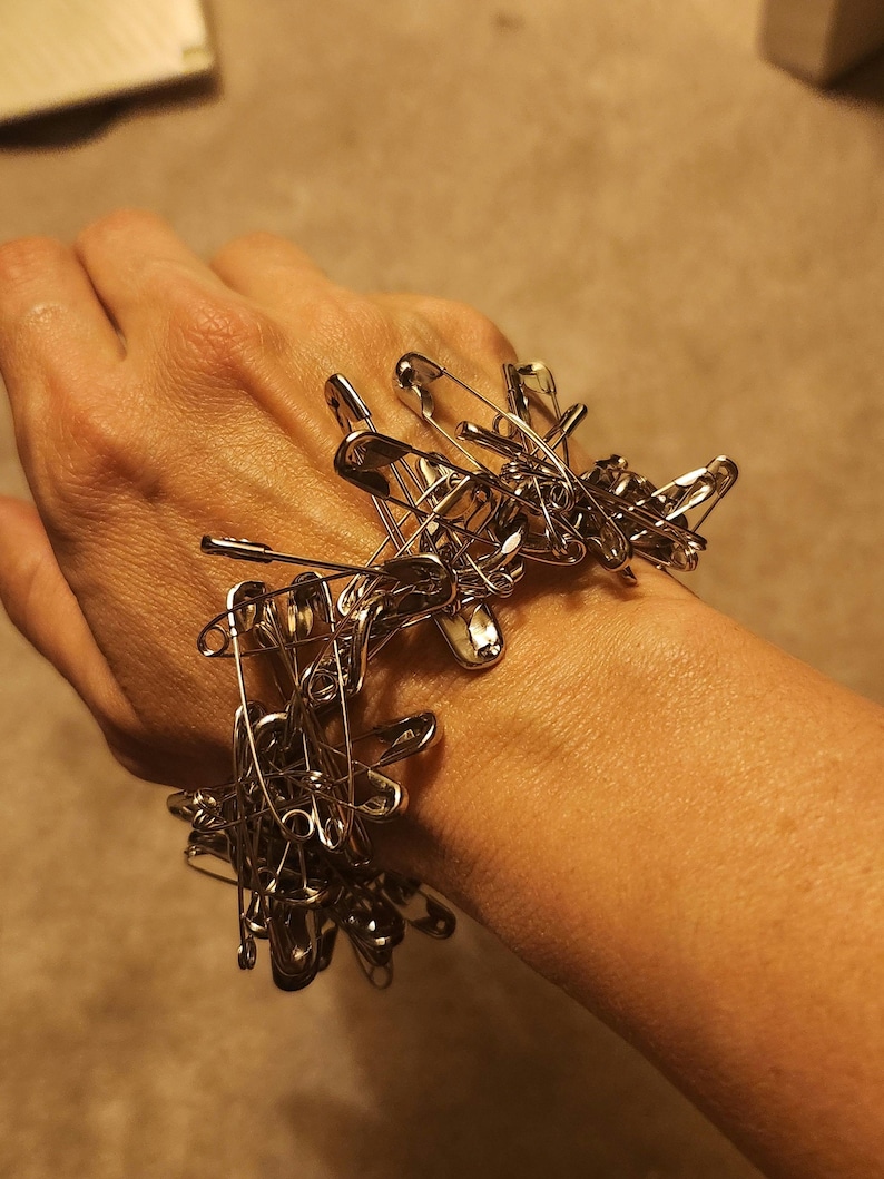 May include: A silver safety pin bracelet made of multiple safety pins linked together. The bracelet is worn on a person's wrist.