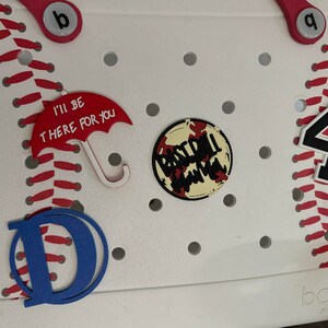 May include: White plastic storage bin with a baseball theme. The bin has red stitching around the edges and a red and white baseball graphic. The bin also has a red umbrella charm with the text "I'll be there for you", a blue letter "D", a baseball-themed charm with the text "Baseball Mama", and a black number "4" charm.