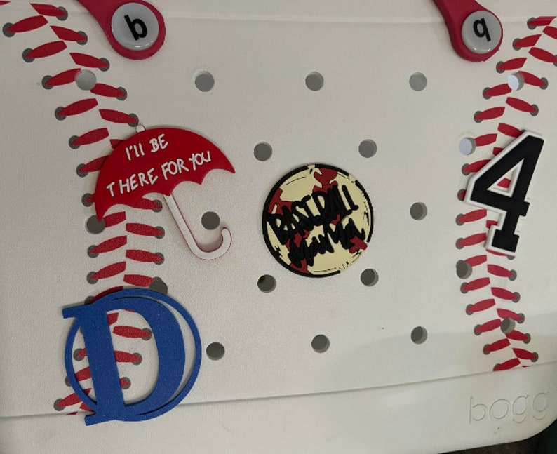 May include: White plastic storage bin with a baseball theme. The bin has red stitching around the edges and a red and white baseball graphic. The bin also has a red umbrella charm with the text "I'll be there for you", a blue letter "D", a baseball-themed charm with the text "Baseball Mama", and a black number "4" charm.