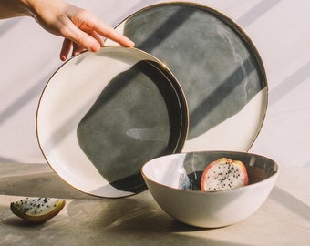 May include: A set of three ceramic plates with a black and white design. The plates have a gold rim and are stacked on top of each other. A bowl with a similar design is in the foreground. A piece of dragon fruit is in the bowl.