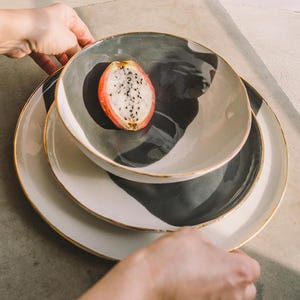 May include: Three ceramic plates with a black and white design and gold trim. The top plate has a slice of dragon fruit in the centre.