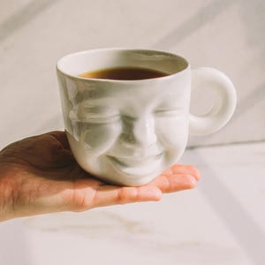 May include: A white ceramic mug with a smiling face design, filled with a dark liquid. The mug has a rounded handle and is held in a person's hand. The mug is on a white surface.