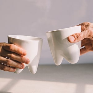 May include: Two white ceramic cups shaped like teeth, held by two hands, with one hand raised in a toast.