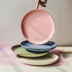 以下が含まれることがあります： ピンク、ブルー、グリーン、ホワイトなど、さまざまな色のセラミックプレート4枚が積み重ねられています。プレートは金色の縁取りがされており、互いに積み重ねられています。一番上のプレートは手で持たれています。