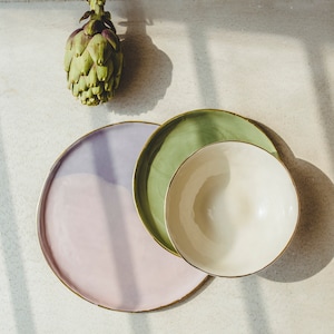 Puede incluir: Tres platos de cerámica con bordes dorados. El plato más grande es rosa claro, el plato del medio es verde y el plato más pequeño es blanco. Los platos están apilados uno encima del otro.