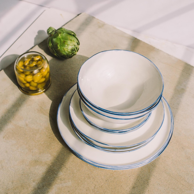 Puede incluir: Un juego de platos de cer&aacute;mica blanca con ribete azul, apilados uno encima del otro. Los platos est&aacute;n sobre una superficie marr&oacute;n claro.