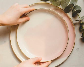 Puede incluir: Dos platos de cerámica rosa con borde dorado, uno ligeramente más pequeño que el otro, están apilados uno encima del otro.