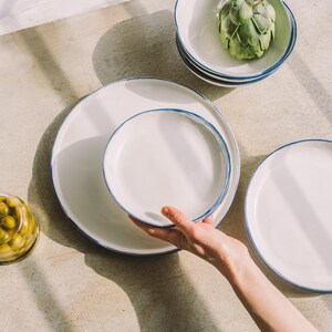 Puede incluir: Un juego de platos de cer&aacute;mica blanca con un borde azul. Los platos est&aacute;n apilados uno encima del otro. Los platos est&aacute;n sobre una superficie gris claro.
