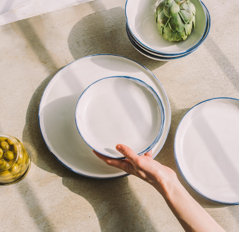 Puede incluir: Un juego de platos de cer&aacute;mica blanca con un borde azul. Los platos est&aacute;n apilados uno encima del otro. Los platos est&aacute;n sobre una superficie gris claro.