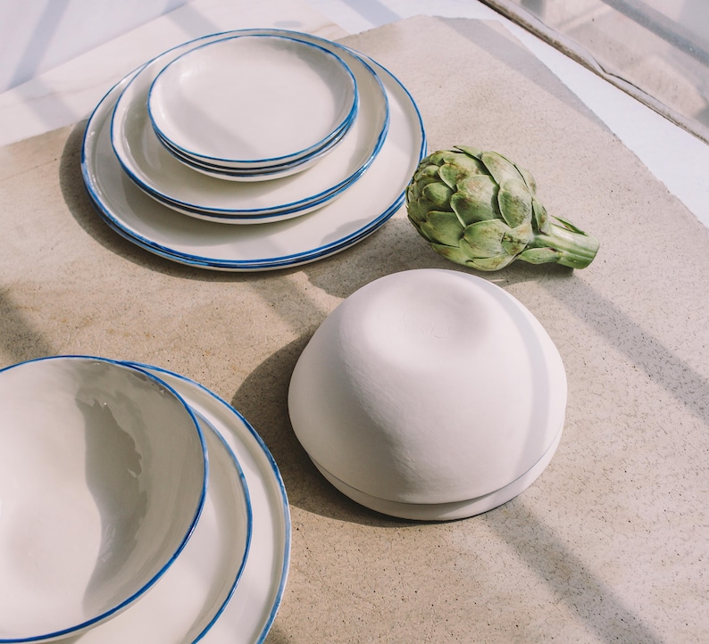 Puede incluir: Un juego de tres platos de cer&aacute;mica blanca con borde azul, un taz&oacute;n de cer&aacute;mica blanca y una alcachofa verde sobre una superficie beige.