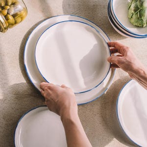 Puede incluir: Dos platos de cer&aacute;mica blanca con un borde azul, uno apilado sobre el otro. Los platos est&aacute;n sobre una superficie marr&oacute;n claro.