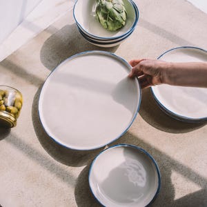 Puede incluir: Un juego de cuatro platos de cer&aacute;mica blanca con borde azul. Los platos est&aacute;n dispuestos en c&iacute;rculo sobre una superficie marr&oacute;n claro. El plato m&aacute;s grande est&aacute; en el centro y el m&aacute;s peque&ntilde;o a la derecha. Hay un tarro de aceitunas en la esquina inferior izquierda de la imagen.