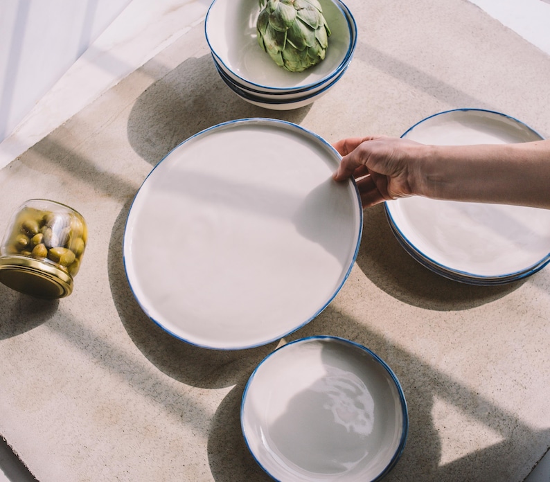 Puede incluir: Un juego de cuatro platos de cer&aacute;mica blanca con borde azul. Los platos est&aacute;n dispuestos en c&iacute;rculo sobre una superficie marr&oacute;n claro. El plato m&aacute;s grande est&aacute; en el centro y el m&aacute;s peque&ntilde;o a la derecha. Hay un tarro de aceitunas en la esquina inferior izquierda de la imagen.