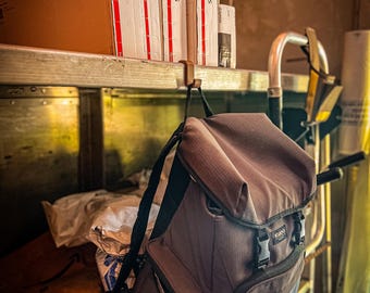 Op de afbeelding: Een bruine rugzak met zwarte banden en gespen hangt aan een haak aan een plank. De rugzak heeft het woord "KODIAK" op de voorkant gedrukt. Verschillende kartonnen dozen met het label "Express" zijn gestapeld op de plank boven de rugzak.