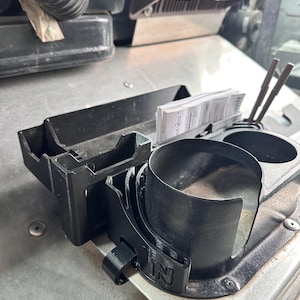 May include: A black plastic vehicle console organizer featuring a rectangular storage compartment, two circular cup holders, and a small slot containing white paper slips. The front of the cup holder section displays a molded letter N logo.