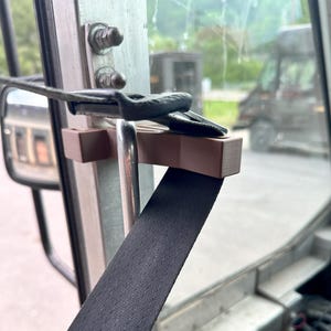 Op de afbeelding: Een close-up van een bruin plastic veiligheidsapparaat voor een veiligheidsgordel. Het apparaat is bevestigd aan een metalen paal en heeft een zwarte band die er doorheen loopt.