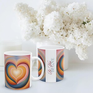 Peut inclure: Deux tasses en céramique blanche avec des motifs abstraits colorés. Une tasse présente une forme de cœur avec un centre blanc et le texte "Be You Be Love".